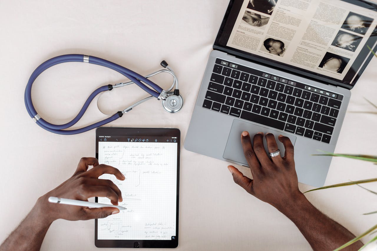 Healthcare professional using tablet and laptop for research with stethoscope on desk.